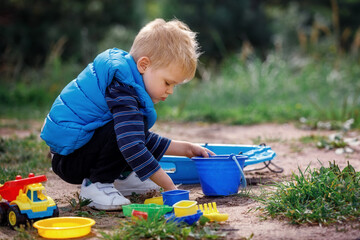 Child squat on the ground playing with sand toys. The child digs the sand into a plastic bucket. Summer outdoor activity for kids. Leisure Time.