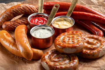 A mix of grilled sausages - Bavarian, round, Cumberland, bratwurst with ketchup and sauce on a wooden table. Traditional Bavarian Beer Snack, close up