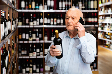 Happy positive senior man talking on mobile phone and choosing bottle of wine at the liquor store
