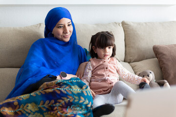 Young Muslim woman with her young daughter at home. Single parent family watching cartoons on...