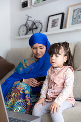 Little toddler watching cartoons on laptop computer. Mother and daughter together. Muslim single parent family at home.