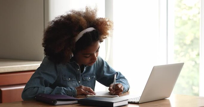 Busy African teen female high school pupil college student sit at table by laptop in headphones listen to video lesson at home write up information. Gen z lady prepare essay homework using modern tech