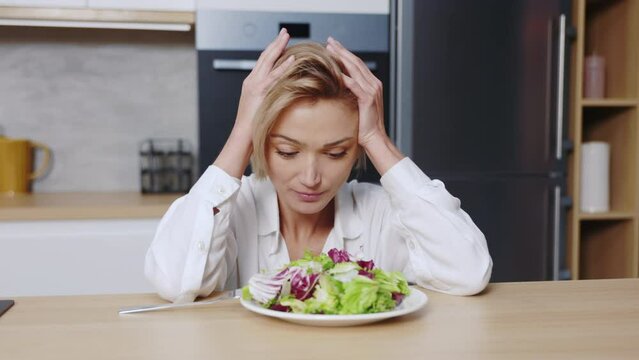 Footage of sad young Caucasian woman with blonde hair sitting at table with plate of salad, holding head in cozy kitchen. Unhappy vegetarian woman in diet. Negative emotions, facial expressions.