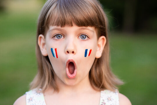 Portrait of surprised 7 years old child with painted cheeks in flag of France walking outdoor. Cute little girl screaming with opened mouth and crazy expression looking at camera. 