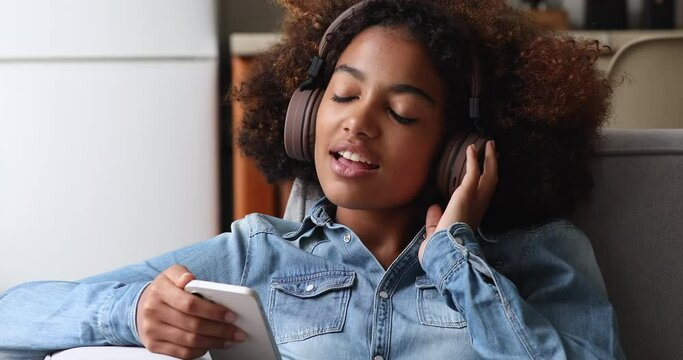 Excited Afro American female teenager relax at home wear wireless headphones listen to music using player app on smartphone. Serene gen z woman relieve stress enjoy favorite melodies with closed eyes