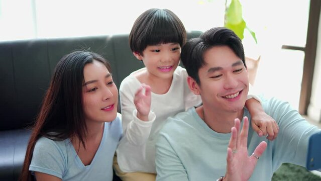 Happy family time, Taking selfie or video call mobile phone of Asian parents dad, mom, newborn baby together sitting on sofa in home, laugh smile. Concept of technology, lifestyle family parenthood.