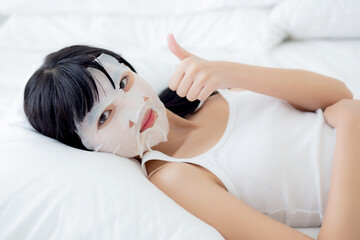 Beautiful young asian woman with sheet facial mask and thumbs up on bed at bedroom, beauty girl...