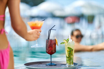 Two fresh cool cocktails standing on round tray near swimming pool. Crop view of female hand...