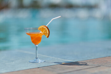 Tasty orange cocktail in glass located on grey tile edge of swimming pool in sunny afternoon....