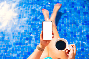 Woman holding mobile phone while sitting by the pool on vacation, blank white screen mockup