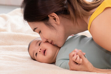 Slective focus Beautiful mom kissing on the cheek newborn with love and caring, Mother showing love to toddler with kiss on cheek. infant baby lying on bed with asian mom.