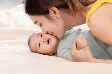 Slective focus Beautiful mom kissing on the cheek newborn with love and caring, Mother showing love to toddler with kiss on cheek. infant baby lying on bed with asian mom.