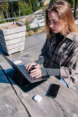 A young, slender girl in a plaid shirt works at a laptop in nature against the backdrop of mountains
