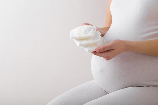 Young adult pregnant woman with big belly isolated on light gray background. Hands holding and showing new white soft baby diapers. Closeup. Empty place for text. Side view.