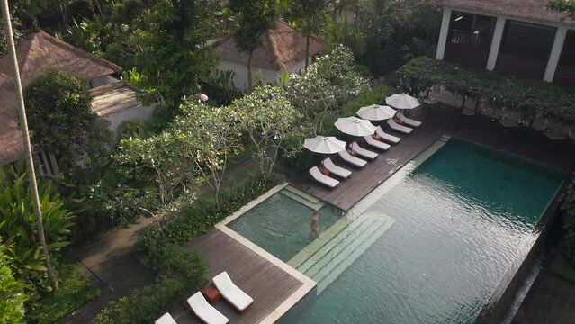 Woman in bikini stepping into luxury swimming pool at tropical jungle resort, dolly out, aerial
