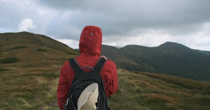 back view, tourist person in a red jacket with a hood and a backpack, looks at the mountains and clouds. Medium shot, daytime, usually spring summer. Carpathians, Ukraine, Europe