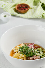 Traditional russian soup - okroshka with fresh vegetables, meat and kvass. Cold summer soup on white table. Aesthetic composition with traditional russian food and spices, textile, spoon.