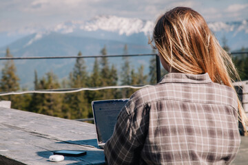 Fototapeta premium A young, beautiful girl in a plaid shirt works on a laptop at a wooden old table in nature against the backdrop of mountains, next to a smartphone and headphones. View from the back.