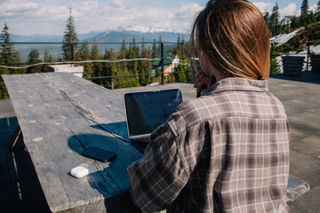 A young, beautiful girl in a plaid shirt works on a laptop at a wooden old table in nature against the backdrop of mountains, next to a smartphone and headphones. View from the back.