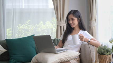Obraz premium Casual young asian woman resting on couch and using laptop, surfing internet, checking email