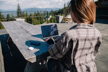 A young, beautiful girl in a plaid shirt works on a laptop at a wooden old table in nature against the backdrop of mountains, next to a smartphone and headphones. View from the back.