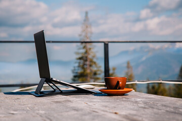 A laptop on a stand, a mouse and a cup of coffee stand on an old wooden table in nature against the backdrop of mountains in clear weather.