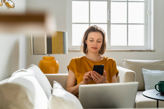 Freelancer using smart phone sitting with laptop on sofa at home