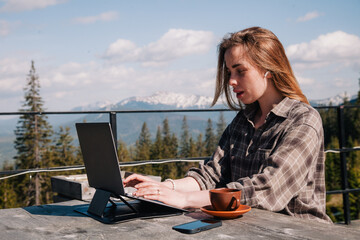 A young, beautiful girl with long hair in a plaid shirt sits at an old wooden table in nature and drinks coffee and works at a laptop against the backdrop of mountains.