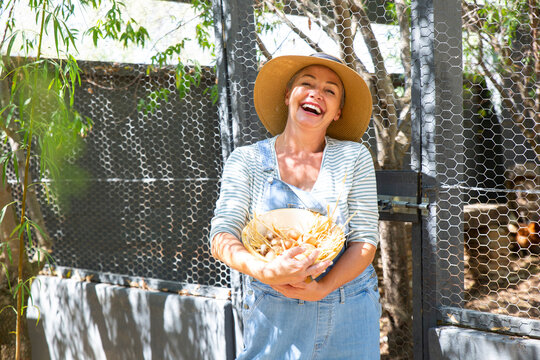 Happy farmer wearing hat holding egg's bowl in front of gate on sunny day