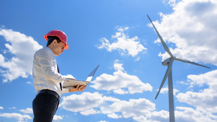 Young man engineer in white shirt and red safety helmet holding a computer and verifying the wind turbine windmill.