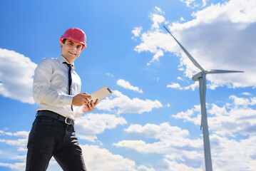 Young man engineer in white shirt and red safety helmet holding a tablet and verifying the wind turbine windmill.