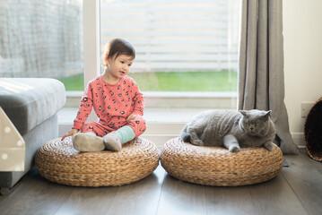 A toddler wearing pink pyjamas, long socks and slippers sits on a wicker stool and looks at her British Short Hair cat sat next to a her in a house in Edinburgh, Scotland, UK