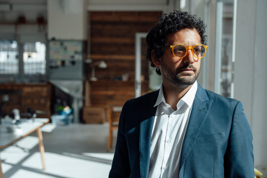 Thoughtful businessman wearing eyeglasses standing in office - Powered by Adobe