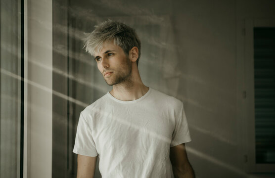 Sad young man with gray hair standing by window