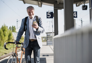 Smiling businessman using smart phone walking with bicycle on platform