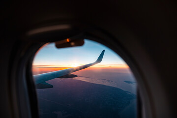 Wing of airplane flying against setting sun seen through porthole