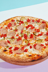 Italian pizza with ham and pickled cucumber on coloured background. Meat pizza with ham and vegetables in minimal style on light blue color. American pizza delivery concept with color backdrop