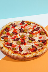 Italian pizza with sausage, jalapeno, ham and paprika on coloured background. Spicy pizza with jalapeno in minimal style on blue and orange color. American pizza delivery concept with color backdrop.