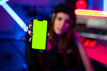 Beautiful caucasian girl in stylish clothes holding a phone with a green screen on a background of neon light in the recording studio