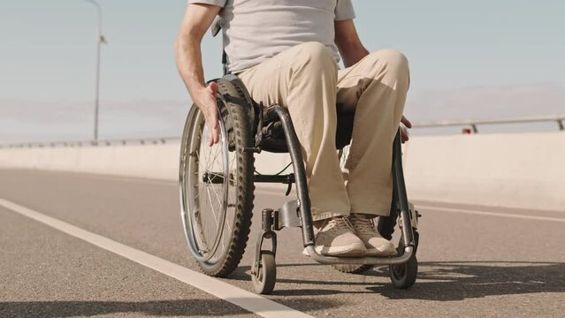 Tilt up or mature Caucasian man with disability riding wheelchair along bridge on sunny day and taking picture of beautiful landscape on smartphone
