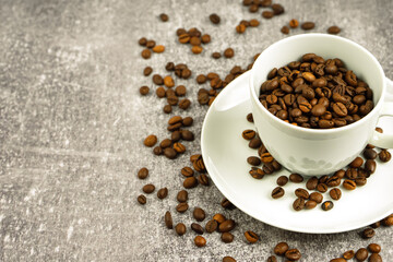White cup of coffee with coffee beans on gray concrete background with scattering of coffee beans. Arabica, Robusta. Drink for office and home. Background for site with copy space for text