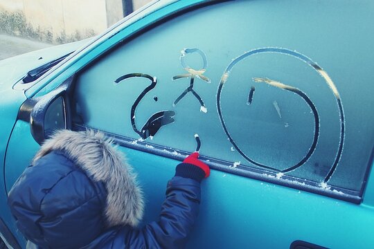 Un ni&ntilde;o haciendo dibujos con el dedo en la ventana de un coche. Ni&ntilde;o con abrigo haciendo monigotes con la escarcha formada sobre el cristal en una fr&iacute;a ma&ntilde;ana de invierno.