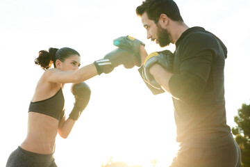 Couple boxing against sky in sunlight. Woman punching man with fist in boxing gloves.