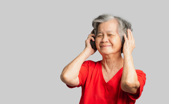 Cheerful elderly Asian woman wireless headphones to listen to a favorite song with a smile while standing on a gray background.