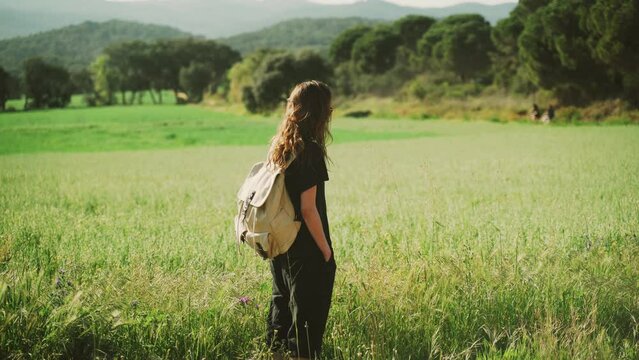 Hiking in the countryside. Young woman with a backpack looking on a flowering field. Girl with long hair in black clothes enjoys a warm sunny day. Travel and recreation. Vintage rucksack. 