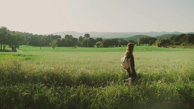 Hiking in the countryside. Young woman with a backpack walks along a flowering field. Girl with long hair in black clothes enjoys a warm sunny day. Travel and recreation. Vintage rucksack.