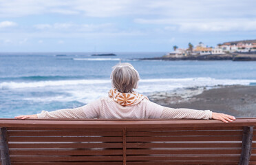 Rear view of senior woman sitting on outdoors bench at sea looking at horizon enjoying freedom and vacation. Active caucasian elderly woman relaxing on a sunny day close to the beach
