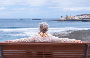 Rear view of senior woman sitting on outdoors bench at sea looking at horizon enjoying freedom and...
