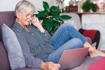Relaxed elderly woman working on laptop at home sitting on sofa in living room. Modern senior lady...