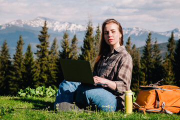 A young, slender girl with loose hair in a plaid shirt and jeans with an orange backpack works on a laptop sitting on green grass against the backdrop of mountains.